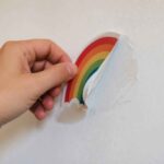 Dłoń odkleja tęczową naklejkę z białej ściany